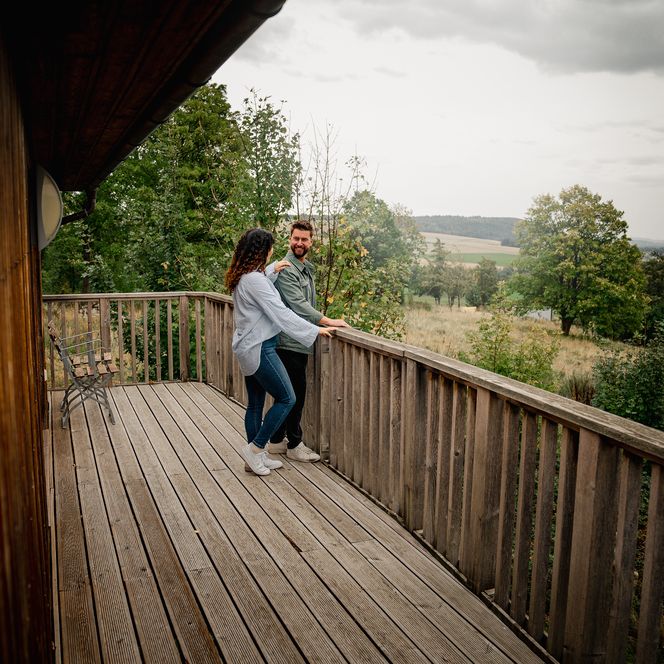 Ein Paar steht auf dem Balkon des Stelzenhauses und blickt über die weite, grüne Landschaft.