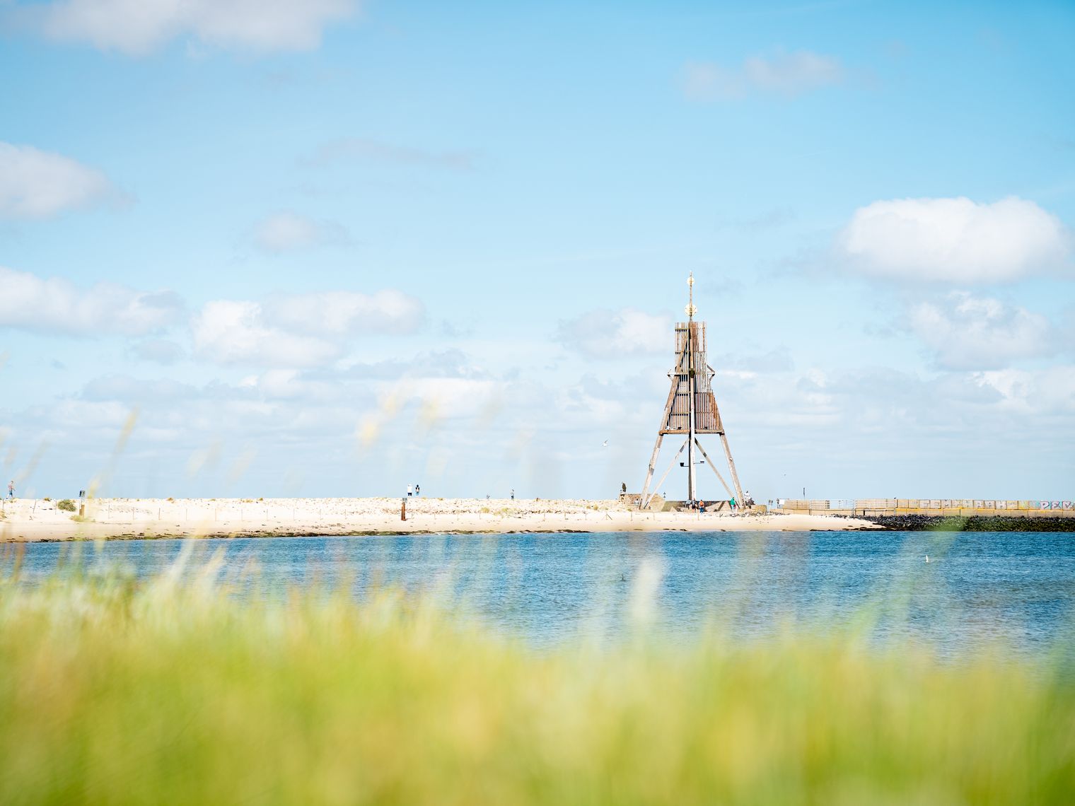 Hölzerne Kugelbake am Strand von Cuxhaven hinter blauem Meer und grünem Dünengras.