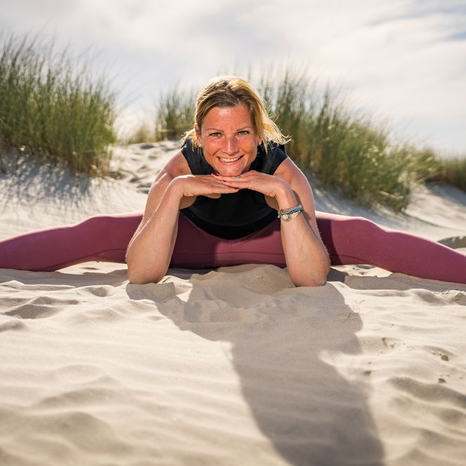 Frau sitzt lächelnd in Yogapose im Sand vor Dünen auf Borkum