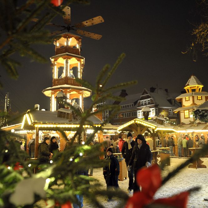 Beleuchtete Weihnachtspyramide mit Marktständen und Besuchern auf einem verschneiten Platz bei Nacht.