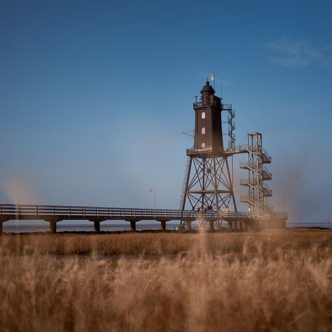 Dunkler Leuchtturm Obereversand mit Steg im Wattenmeer an der Wurster Nordseeküste bei blauem Himmel.