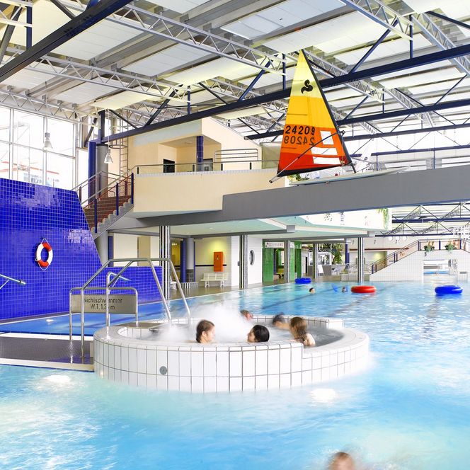 Innenansicht eines modernen Hallenbads mit blau gefliesten Wänden, Whirlpool und großem Becken.
