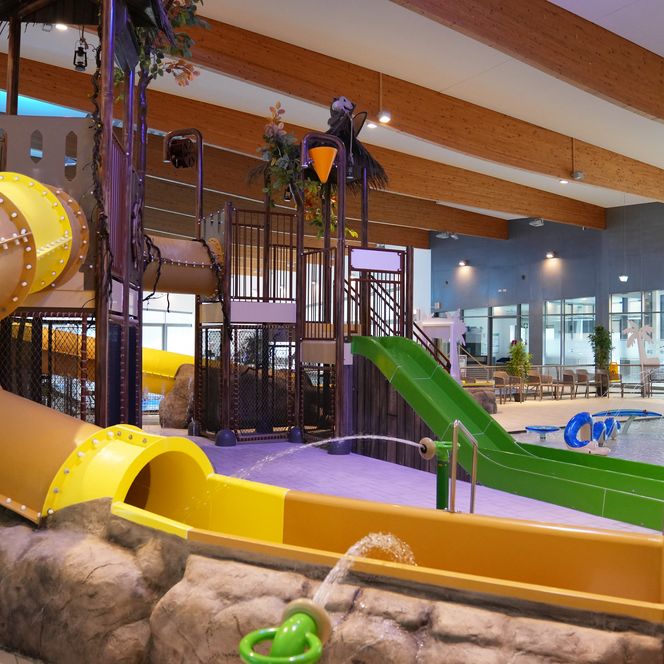 Ein modernes Hallenbad mit Rutschen und Wasserbereich. Holzdecken und Pflanzen schaffen eine freundliche Atmosphäre.