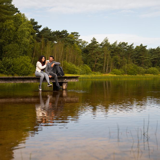 Ein Paar sitzt auf einem Holzsteg am Großen Bullensee, umgeben von Bäumen und klarem Wasser.