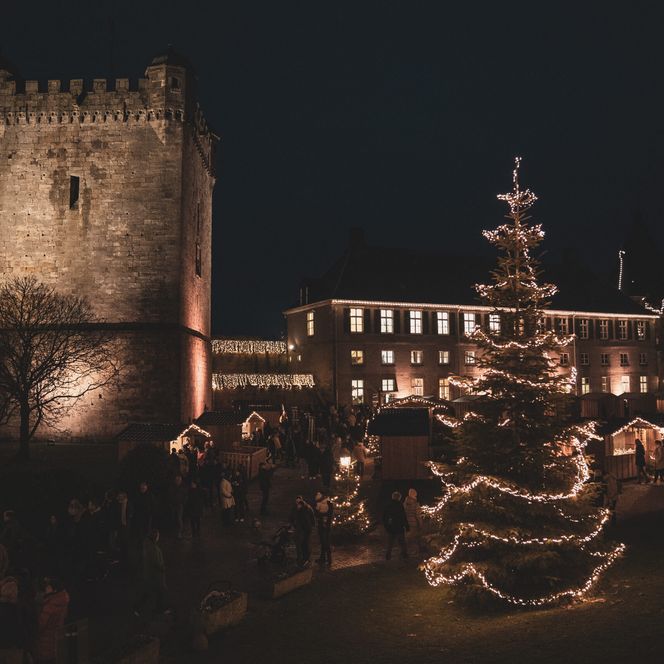 Beleuchtete Burg Bentheim mit Weihnachtsmarktständen und festlich geschmücktem Tannenbaum bei Nacht.