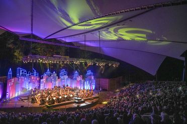 Orchester spielt vor farbig beleuchteter Kulisse auf der Freilichtbühne Meppen unter Zeltdach.