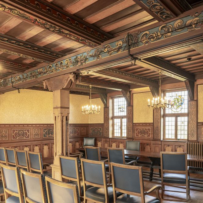 Historischer Gerichtssaal mit bemalter Holzbalkendecke, Kronleuchtern und dunklem Mobiliar.