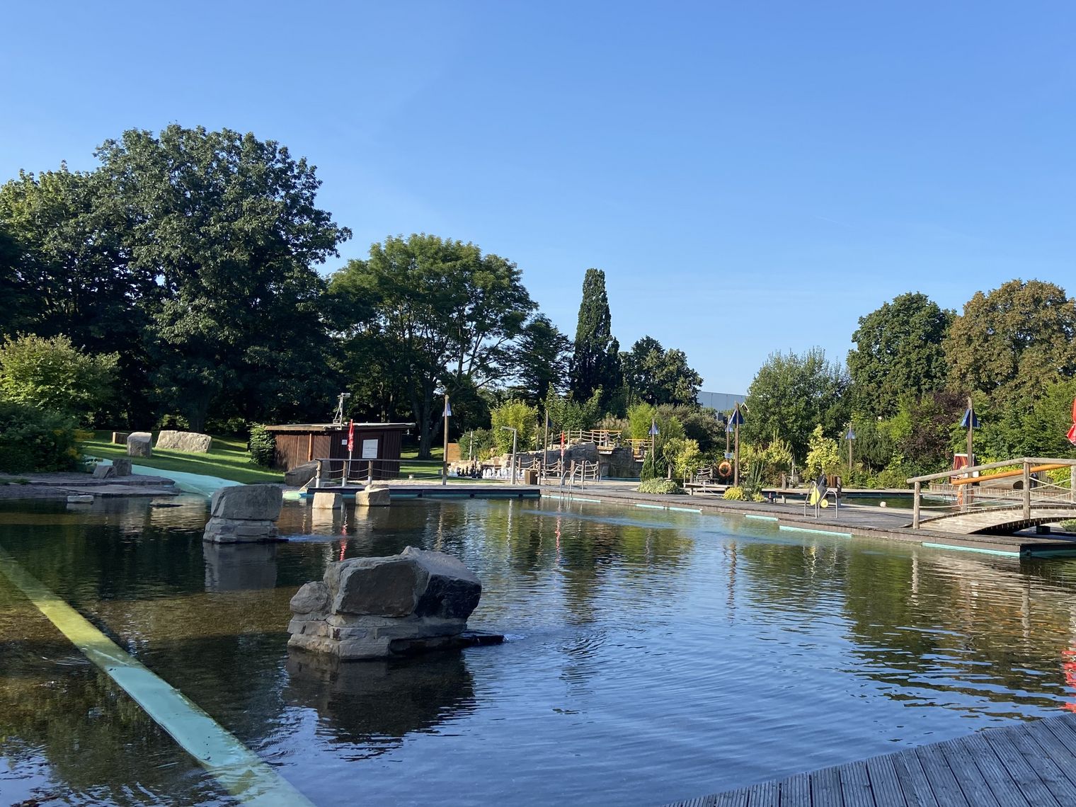 Ein weitläufiger Park mit Teich, Holzsteg und Brücken, umgeben von vielfältigen grünen Bäumen.