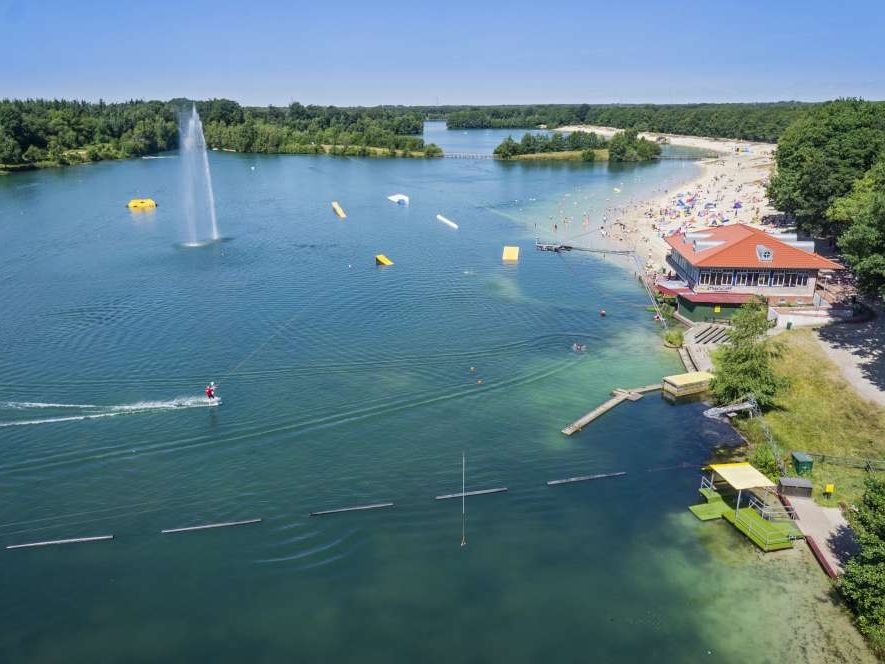 Weitläufiger Badesee mit Sandstrand, Wassersportanlagen, Wakeboardbahn und umgebendem Wald.