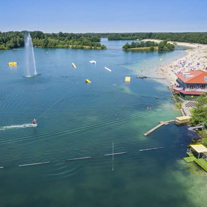 Weitläufiger Badesee mit Sandstrand, Wassersportanlagen, Wakeboardbahn und umgebendem Wald.