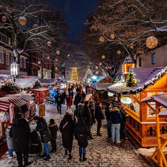 Beleuchtete Buden auf dem verschneiten Weihnachtsmarkt in Stade, Menschen schlendern durch den Schnee.