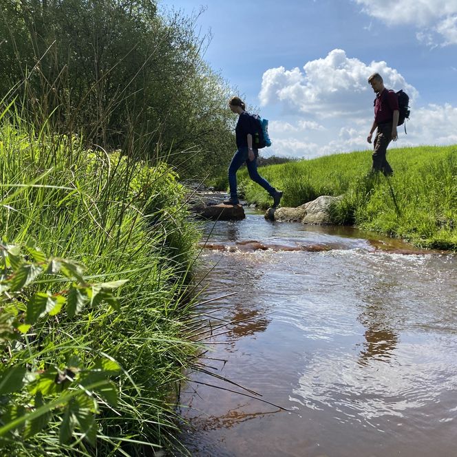 Zwei Wandernde überqueren auf Steinen einen Bach im grünen Ohetal unter blauem Himmel.