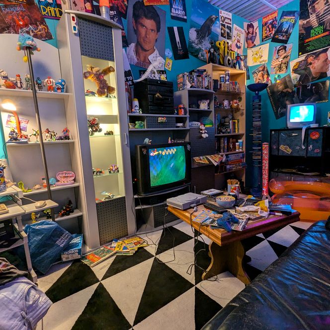 Das Bild zeigt einen Raum voller Retro-Spielzeug und Spielekonsolen, umgeben von bunten Postern.