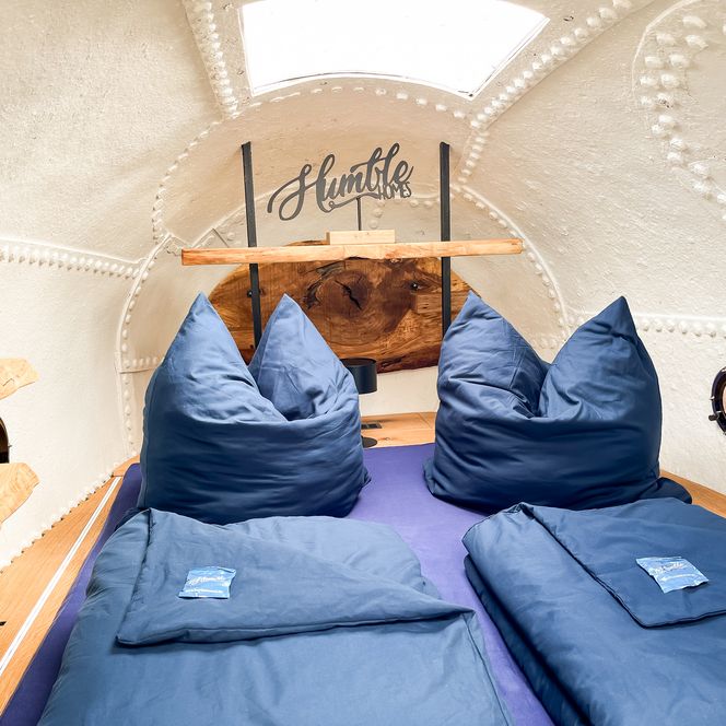 Runde weiße Schlafkabine mit Nieten, Dachfenster, Bullauge und blau bezogenem Bett.