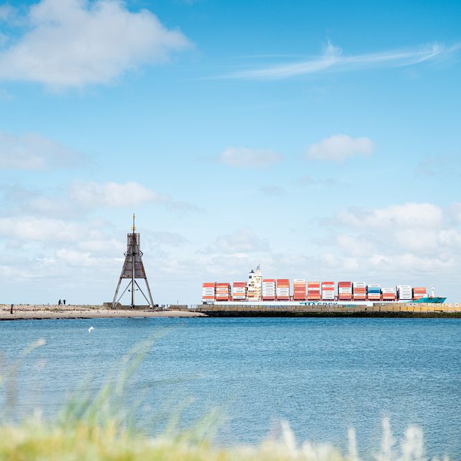Die Kugelbake in Cuxhaven am Wasser, im Hintergrund fährt ein großes Containerschiff vorbei.