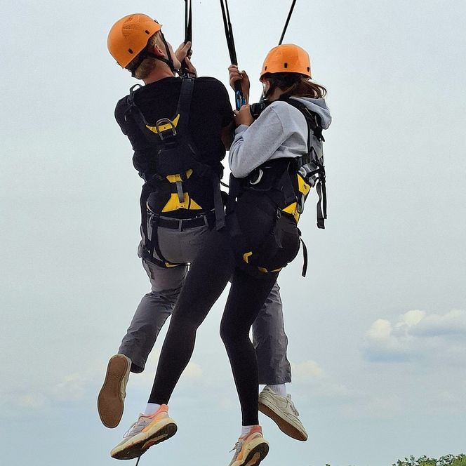 Zwei Personen mit Klettergurten und Helmen schweben an Seilen über Baumwipfeln.