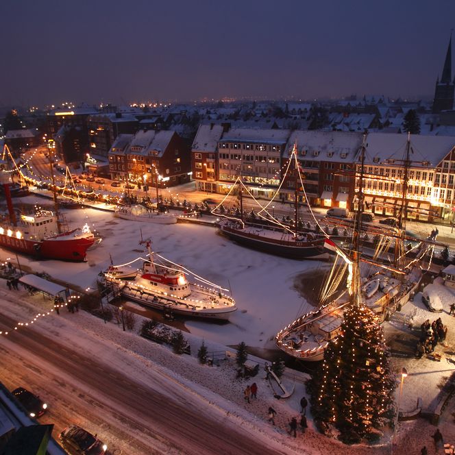 Verschneiter Hafen mit hell erleuchteten Schiffen, Weihnachtsbaum und festlich geschmückten Häusern.