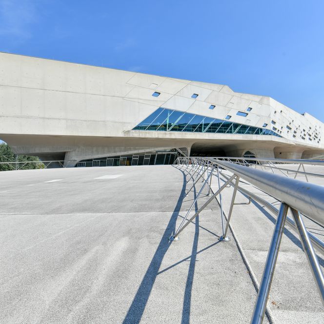 Modernes Betongebäude mit futuristischer Form und schrägen Glasflächen unter blauem Himmel.