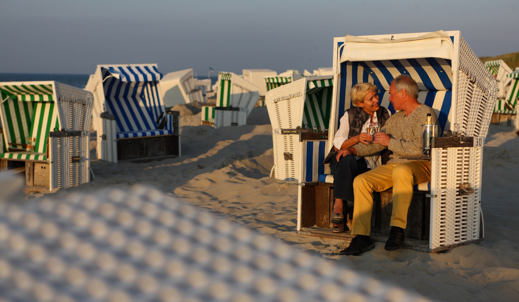 Ein älteres Paar sitzt in einem Strandkorb und stößt mit Sekt an., © Kurverwaltung Wangerooge