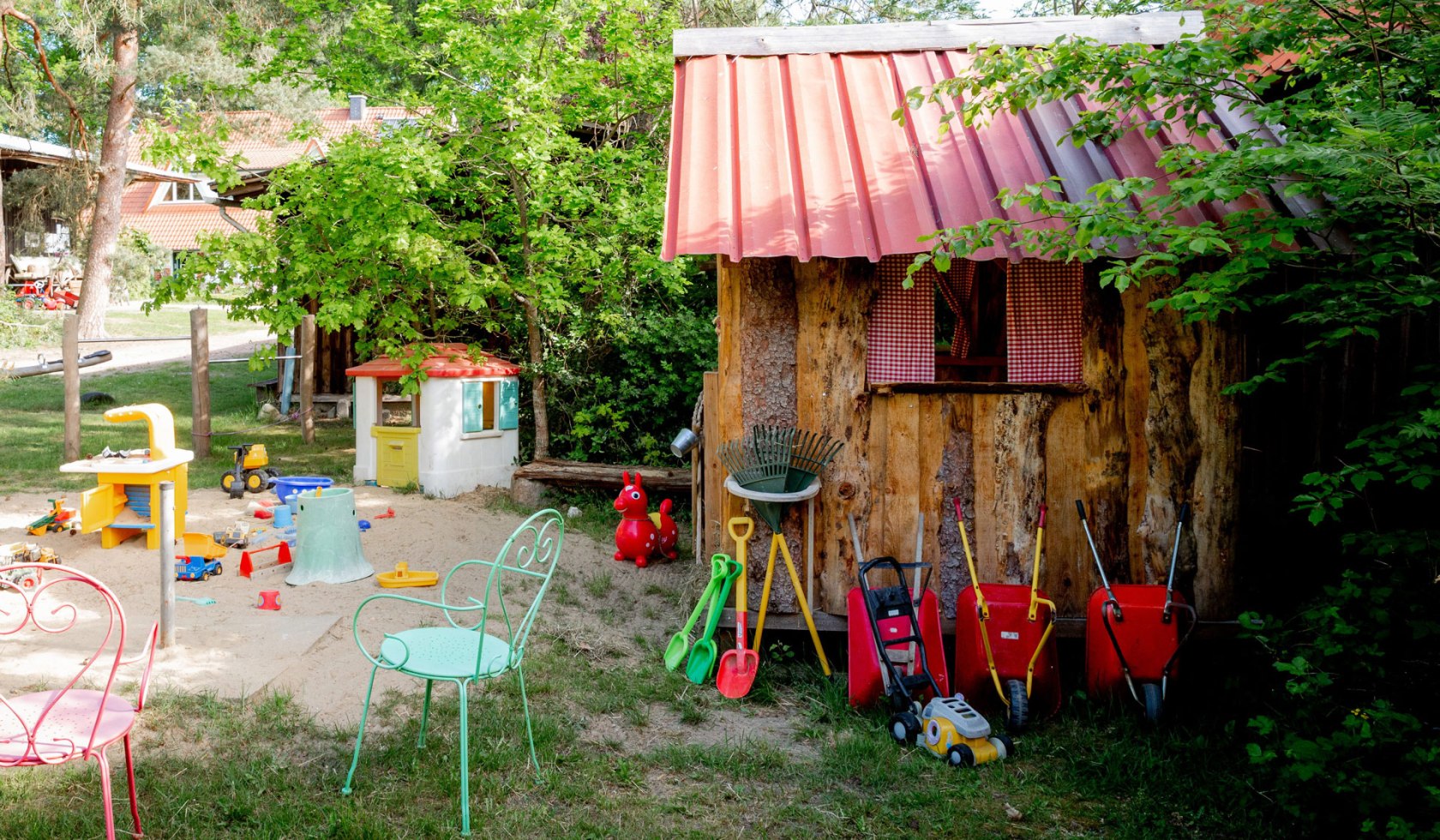Eine kleine Holzhütte für Kinder steht im Grünen. Davor befindet sich eine Sandfläche mit Stühlen und vielen Spielzeugen., © Claudia Drewes / Christina Opeldus