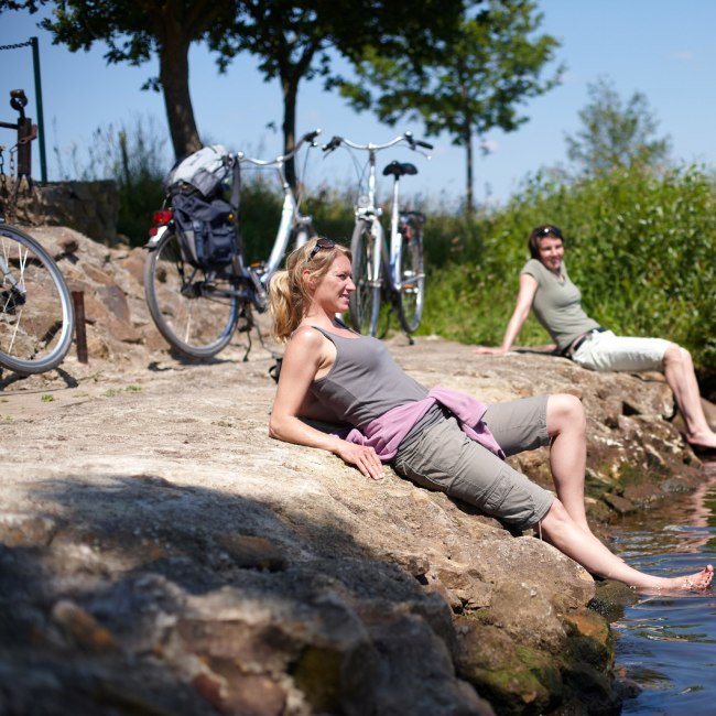 Radfahrerinnen sitzen an der Ems und halte ihre Füße ins Wasser., © Emsland Touristik GmbH