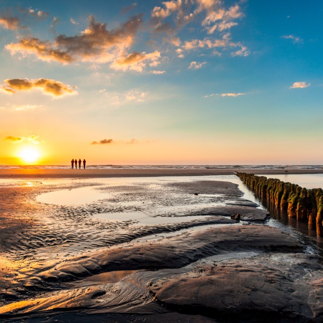 Drei Personen stehen am Nordstrand von Norderney und beobachten den Sonnenuntergang, © TMN/Janis Meyer 